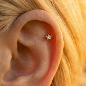 Könnte beinhalten: Nahaufnahme eines kleinen, sternförmigen Ohrrings mit einem klaren Edelstein, der in einem goldfarbenen Metall gefasst ist. Der Ohrring befindet sich am oberen Ohrknorpel. Der Hintergrund ist verschwommen und hat einen warmen, goldenen Farbton.