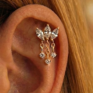 May include: A gold-coloured ear piercing. It features three marquise-cut clear gemstones in a floral arrangement. Below, three smaller round clear gemstones dangle from chains, adding movement and sparkle to the design.
