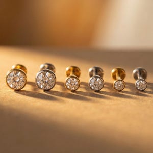 Pode incluir: Uma coleção de seis brincos de pressão com pedras preciosas redondas e transparentes em molduras de ouro e prata. Os brincos estão dispostos em uma fileira, com tamanhos variados, e são exibidos em uma superfície quente e neutra.