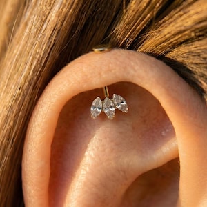 Puede incluir: Un piercing dorado con tres gemas claras de talla marquesa. El piercing está engarzado en una barra curva, con un pequeño perno redondo en la parte superior. La joya está colocada sobre el fondo de una oreja y cabello.