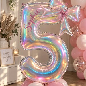 May include: A large iridescent number five foil balloon featuring a prominent bow at the top and the name Sophie printed in pink script. The balloon is anchored by a cluster of light pink balloons and displays a reflective, multi-coloured finish.