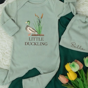 May include: A sage green baby outfit set featuring a ribbed long-sleeved onesie with a duck graphic and the text "LITTLE DUCKLING". The set includes a matching knotted hat with the name "Bella" embroidered on it. The outfit is displayed with sunglasses and flowers.