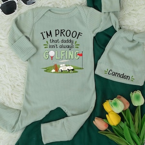 May include: A light green baby onesie with the text "I'M PROOF that daddy isn't always GOLFING" and a golf cart graphic. A matching hat with the name "Camden" is also included. The outfit is ribbed and made of soft material.