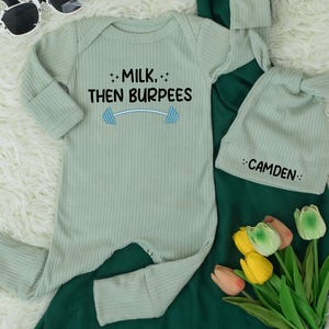 May include: A light green baby onesie with the text "MILK, THEN BURPEES" and a barbell graphic. A matching knotted hat with the name "CAMDEN" is also included. The set is displayed with sunglasses and flowers.