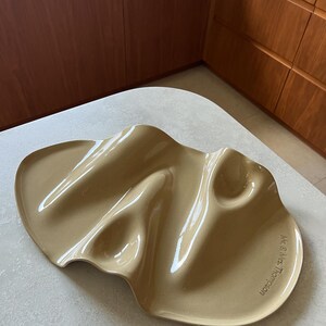 Op de afbeelding: Een beige keramieken serveerblad met een onregelmatige, organische, golvende vorm en een glanzende afwerking. Het oppervlak heeft verschillende verzonken delen en is gegraveerd met de tekst "Mr. & Mrs. Thompson" langs de gebogen rand.