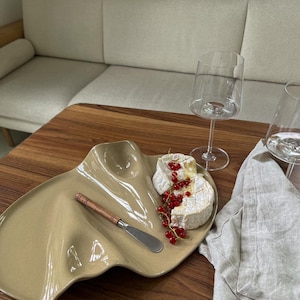Könnte beinhalten: Eine beige Servierplatte aus Keramik mit organischer, gewellter Form. Auf der Platte liegen ein Stück Brie-Käse mit roten Johannisbeeren und ein kleines Käsemesser aus Metall mit Holzgriff. Zwei klare Weingläser stehen daneben.