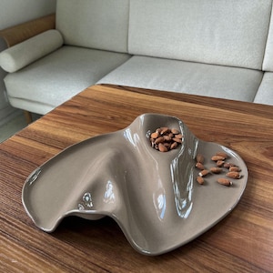 May include: A glossy, taupe-colored ceramic serving dish with an irregular, undulating shape featuring a deep, recessed circular well containing a small pile of brown almonds, with additional almonds scattered across the smooth, curved surface.