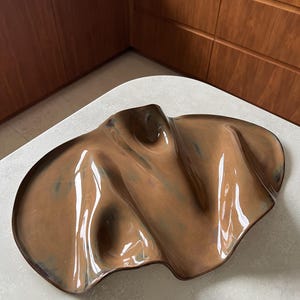 Pode incluir: Uma bandeja de cerâmica grande de formato orgânico irregular com um esmalte brilhante em tons de castanho. A superfície apresenta reentrâncias profundas e cristas elevadas que criam uma aparência fluida e ondulada com tons escuros.