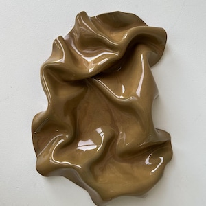 Op de afbeelding: Een glanzend, bruinachtig keramisch dienblad met een onregelmatig, golvend en gevouwen oppervlak. Het object heeft een gladde, reflecterende afwerking met diepe rondingen en verhoogde randen die een vloeiende, organische vorm creëren.