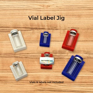 May include: A collection of five plastic vial label jigs in white, blue, and red, each featuring integrated measurement markings. The jigs are designed to hold small glass vials, with one central red jig displaying a label reading Your Peptide Label.