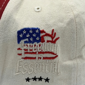 May include: A baseball cap with a cream-colored crown, a red brim, and a blue and red embroidered design. The design includes a stylized American flag and the words "Freedom in Essential". Four black stars are embroidered below the text.