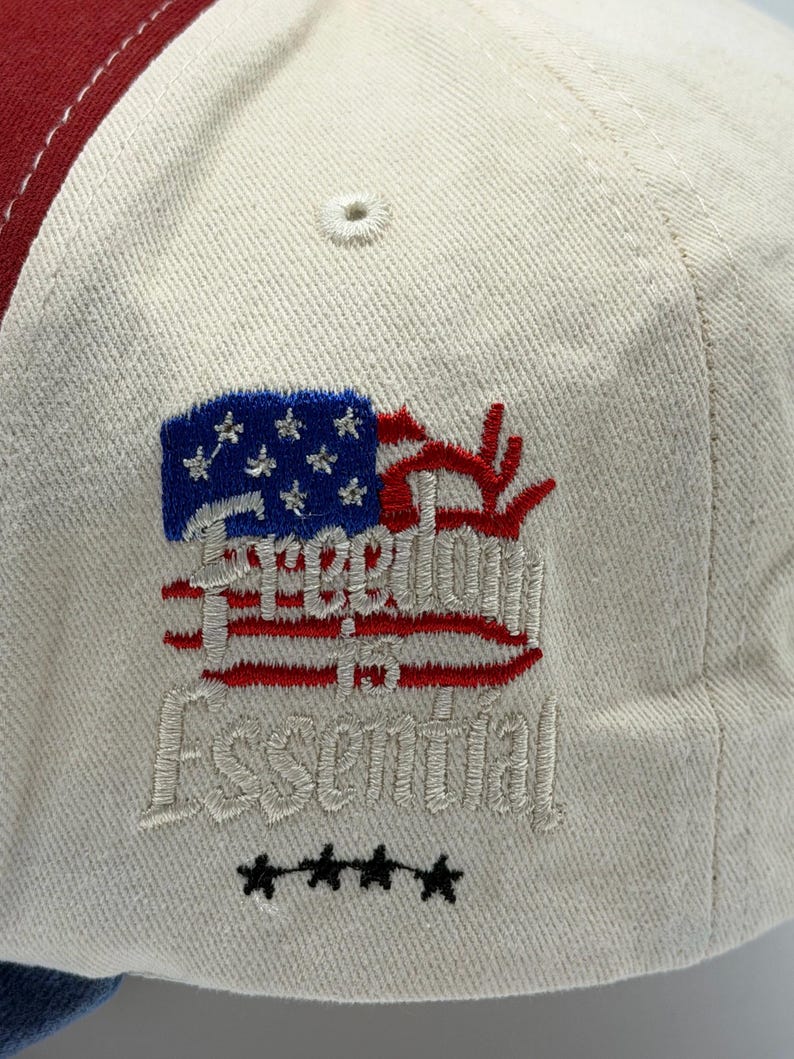 May include: A baseball cap with a cream-colored crown, a red brim, and a blue and red embroidered design. The design includes a stylized American flag and the words "Freedom in Essential". Four black stars are embroidered below the text.