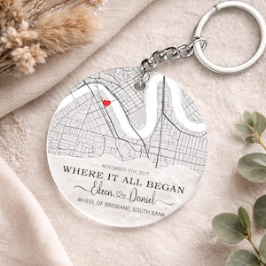 Puede incluir: Un llavero redondo con un diseño de mapa y un corazón rojo que marca una ubicación. El texto "WHERE IT ALL BEGAN" está encima de los nombres "Eileen & Daniel" y la fecha "NOVEMBER 9TH, 2017". También se incluye el texto "WHEEL OF BRISBANE, SOUTH BANK".