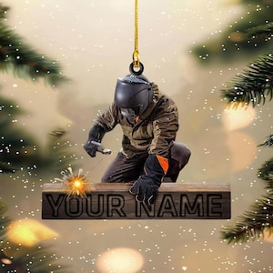 May include: A hanging ornament featuring a welder in a black helmet, brown jacket, and black gloves, working on a wooden beam with the text YOUR NAME engraved on it. The welder is creating a bright orange spark while suspended by a gold cord.