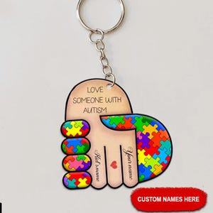 May include: A keychain with a hand design, featuring a puzzle piece pattern in various colors. The text "LOVE SOMEONE WITH AUTISM" is displayed. The keychain includes a metal ring and chain, with space for custom names.