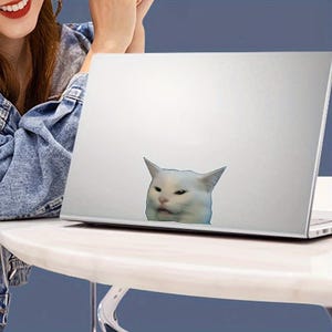 Op de afbeelding: Een witte laptop met een sticker van een witte kat met een chagrijnige uitdrukking. De kattensticker bevindt zich aan de rechteronderkant van de laptop. De laptop staat op een witte tafel.