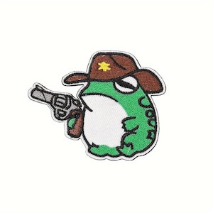 May include: Embroidered patch featuring a green frog wearing a brown cowboy hat with a yellow star and holding a silver revolver. The frog has a white belly and black outlines. The patch is on a white background.