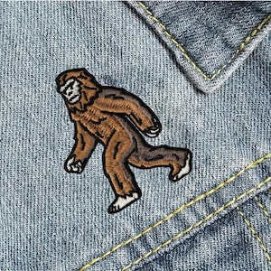 May include: Embroidered patch featuring a brown Bigfoot figure in a walking pose. The patch has a detailed design with black outlines and white accents on the feet and face. The patch is placed on a denim background.