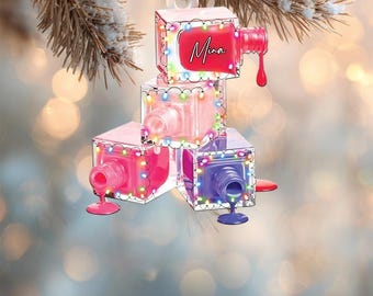 May include: A nail polish bottle ornament with a red bottle and the name "Mina" on the top. The ornament is made of stacked, clear, cube-shaped bottles of pink, red, and purple nail polish, each with a string of lights around it.