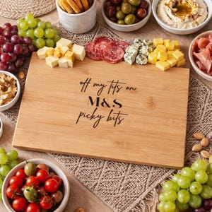 Könnte beinhalten: Ein Holzservierbrett mit dem Text "off my tits on M&S picky bits" ist von verschiedenen Snacks umgeben. Dazu gehören Trauben, Käsewürfel, Oliven, Salami, Hummus und Brotstangen. Ideal für einen gemütlichen Abend.