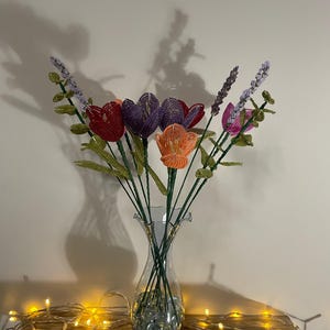 May include: A clear glass vase with a blue base holds a bouquet of beaded flowers. The flowers include red, purple, and orange tulips, and lavender sprigs. The vase sits on a wooden surface, with a string of lights in front.