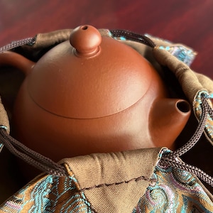 Könnte beinhalten: Eine runde, matte, braune Teekanne aus Ton mit einem kleinen Deckelknauf und einem geschwungenen Ausguss, die in einem Stoffbeutel mit Kordelzug steckt. Der Beutel hat eine beigefarbene Basis mit einem komplexen Webmuster in Türkis, Gold und Lila.