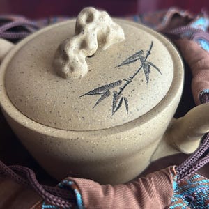 May include: A beige ceramic teapot with a lid featuring a bamboo design and a unique handle. The teapot is nestled in a brown and blue patterned fabric pouch, suggesting a tea set.