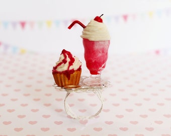 May include: A ring featuring a miniature cupcake and a milkshake. The cupcake has a brown base with white frosting and red drizzle. The milkshake is pink with white whipped cream, a cherry, and a red straw. The ring is silver.