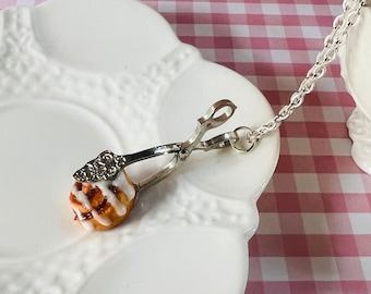 May include: A miniature cinnamon roll held by silver-colored tongs with a decorative handle. The tongs are attached to a chain, resting on a white, scalloped plate. The background features a pink and white checkered pattern.