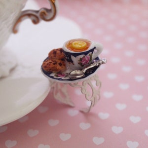 Könnte beinhalten: Ein Miniatur-Teeservice-Ring mit einer weißen Teetasse mit Zitronenscheibe, einem kleinen Teller mit Keksen und einem winzigen Löffel. Die Teetasse und der Teller haben blaue und rosa Blumenmuster. Der Ring ist aus silberfarbenem Metall.