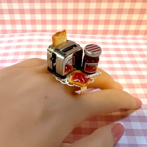 Könnte beinhalten: Ein ausgefallener Ring mit einem Miniatur-Chrom-Toaster, einer Toastscheibe und einem Marmeladenglas. Der Ring hat auch ein Stück Toast mit Marmelade, alles auf einer dekorativen silbernen Basis.