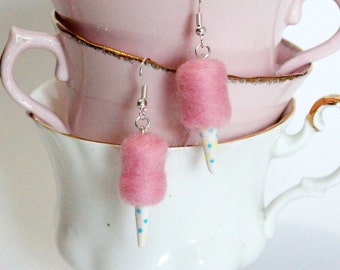 May include: Cotton candy earrings with pink felt candy and white cone-shaped bottoms decorated with blue and yellow dots. The earrings hang from silver-colored hooks. The background includes pink and white teacups.