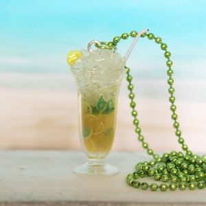 May include: A miniature cocktail pendant necklace featuring a clear glass with ice, lemon slices, and mint. A green beaded chain and a small lemon slice adorn the top. The background is a blurred beach scene.