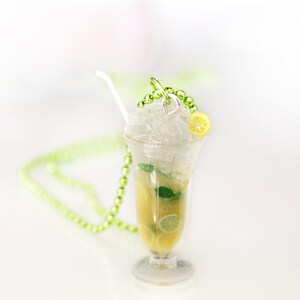 May include: A clear glass pendant necklace shaped like a refreshing cocktail. The pendant features a yellow drink with ice, lemon slices, and mint. A white straw and green beaded chain complete the design. The necklace is on a white surface.