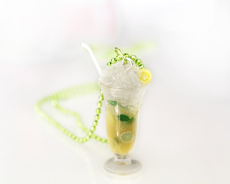 May include: A clear glass pendant necklace shaped like a refreshing cocktail. The pendant features a yellow drink with ice, lemon slices, and mint. A white straw and green beaded chain complete the design. The necklace is on a white surface.