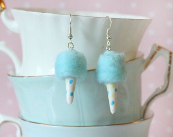 May include: A pair of whimsical earrings shaped like cotton candy. Each earring features a fluffy, light blue cotton candy top and a white cone with blue and yellow polka dots. The earrings hang from silver-colored hooks.