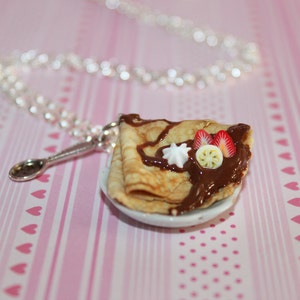 Peut inclure: Un collier argenté avec un pendentif en forme de dessert miniature de crêpe. La crêpe est décorée de sauce au chocolat, de fraises, de crème fouettée et d'une tranche de citron. Le pendentif est posé sur une petite assiette blanche.