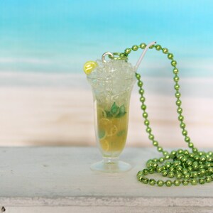May include: A miniature cocktail pendant with a green beaded necklace. The pendant is a clear glass with a yellow-green liquid, ice, and a lemon slice. A white straw and a yellow bead are also visible.
