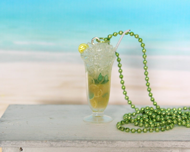 May include: A miniature cocktail pendant with a green beaded necklace. The pendant is a clear glass with a yellow-green liquid, ice, and a lemon slice. A white straw and a yellow bead are also visible.