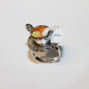以下が含まれることがあります： ミニチュアのティーカップのデザインが施されたシルバーリング。 ティーカップには、茶色の液体、レモンスライス、角砂糖が入っています。 「TEA」という文字の小さなタグがカップからぶら下がっています。 ティーカップはソーサーの上にあります。