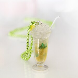May include: A miniature cocktail pendant with a clear glass, filled with a yellow liquid, ice, and a lemon slice. A white straw and green beaded necklace are also present. The pendant is designed to resemble a refreshing drink.