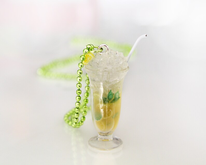 May include: A miniature cocktail pendant with a clear glass, filled with a yellow liquid, ice, and a lemon slice. A white straw and green beaded necklace are also present. The pendant is designed to resemble a refreshing drink.