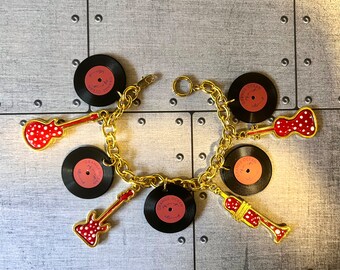 May include: A gold-colored charm bracelet featuring charms of miniature red guitars, trumpets, and vinyl records. The records have a salmon-colored label with the text "ORANGE OCEAN". The guitars and trumpet are red with white polka dots.