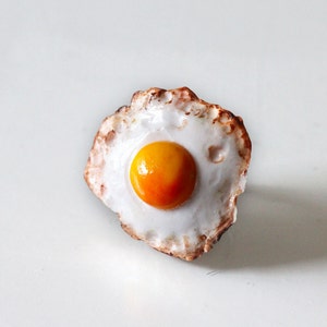 May include: A ceramic fried egg with a bright orange yolk and a white egg white. The egg white has a slightly browned, textured edge. The egg is centered on a white background.