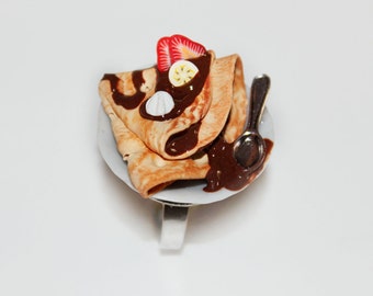 May include: A miniature dessert ring featuring a crepe with chocolate sauce, topped with a strawberry slice, banana, and white candy. The crepe sits on a small white plate with a tiny spoon.