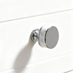 Puede incluir: Primer plano de un armario blanco con dos tiradores de cajón circulares plateados. Los tiradores tienen una superficie brillante y reflectante y están montados sobre una superficie blanca ranurada. El armario parece ser de madera.