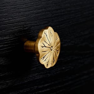 May include: A gold-coloured cabinet knob with a leaf design, set against a black wooden surface. The knob's surface is textured with a central circular detail. The image is a close-up of the knob.