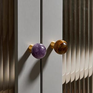 May include: Close-up of a gray cabinet door with two decorative knobs. One knob is a polished purple stone, and the other is a polished brown wood grain. Both knobs have a gold-colored base.