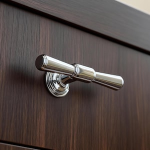May include: A polished chrome cabinet pull handle with a cylindrical design. The handle is mounted on a dark brown wooden cabinet door. The handle has a circular base and a horizontal bar with rounded ends.