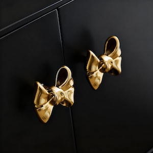 Pode incluir: Close-up de dois puxadores dourados em forma de laço contra uma porta de armário preta. Os puxadores são altamente polidos e refletem a luz, adicionando um toque de elegância. Ferragens decorativas para móveis.